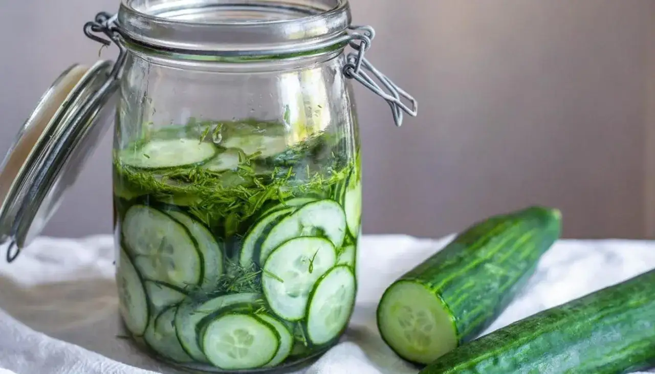 En burk med skivade gurkor och dill, redo att bli picklad gurka. Två hela gurkor ligger bredvid.