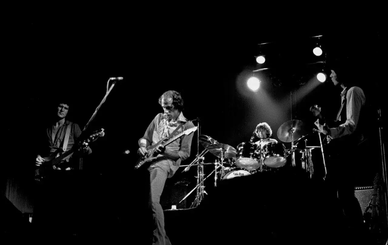 Członkowie zespołu Dire Straits na scenie, grający na gitarach i perkusji.
