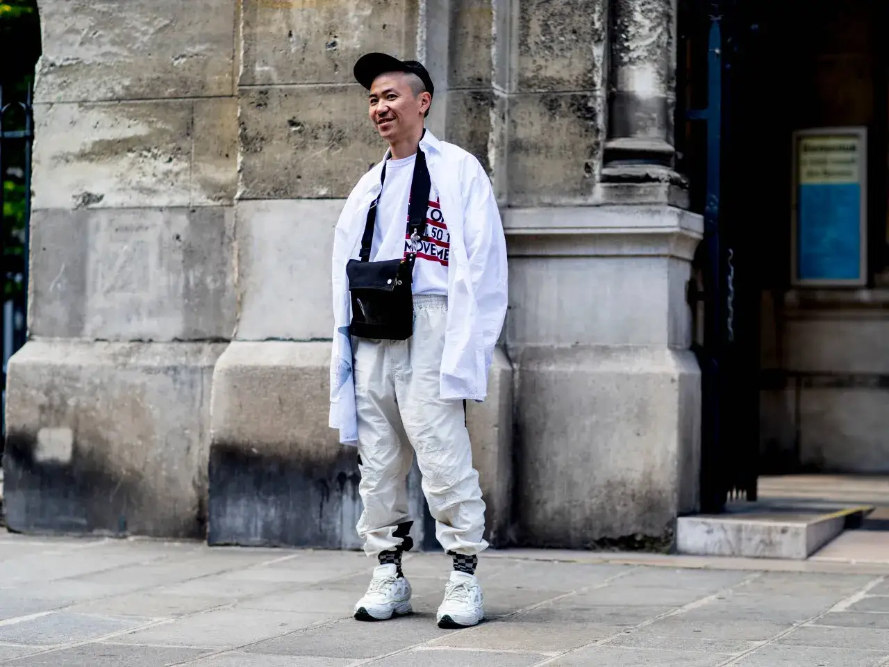 Un homme porte un outfit sneakers homme blanc, un t-shirt graphique et une casquette noire, avec un sac bandoulière noir.