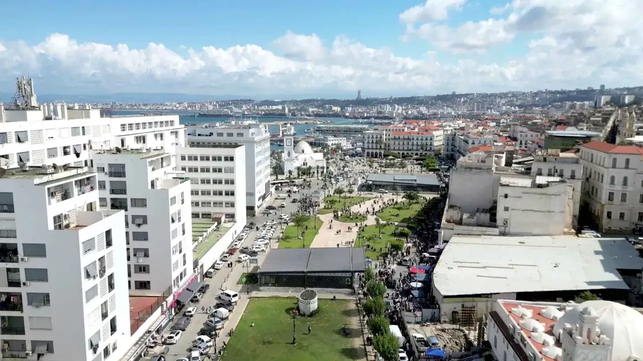 Vue aérienne d'Alger, avec des bâtiments blancs modernes et anciens, un parc verdoyant animé, et la mer en arrière-plan. L'expression de Bab El Oued se lit dans cette scène vibrante.
