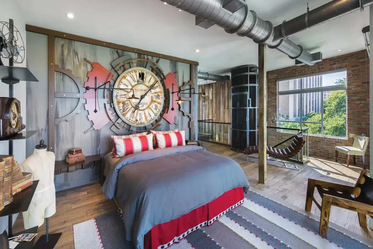Dormitorio con gran reloj de engranajes, ventilador industrial y tuberías visibles. La decoración steampunk crea un ambiente único.