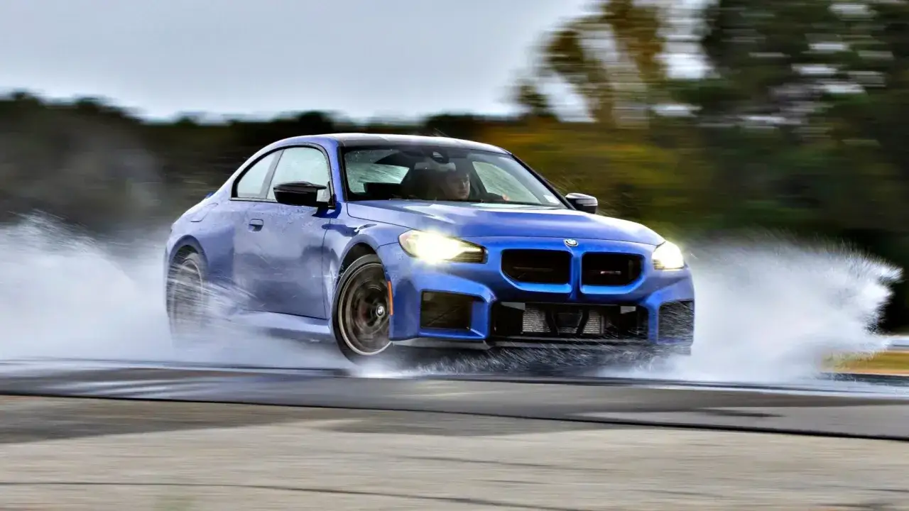 Niebieskie BMW M2 Competition ślizga się po mokrym torze, tworząc fontanny wody.