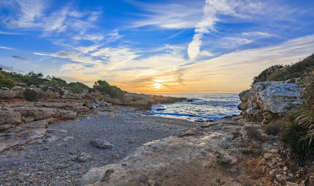 Atardecer en una cala rocosa de Alcossebre. El sol se pone sobre el mar Mediterráneo, pintando el cielo de tonos naranjas y azules. Un lugar perfecto que ver en Alcossebre.
