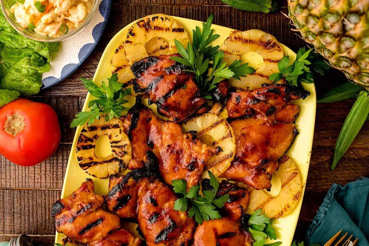 Saftiges Huli Huli Chicken mit gegrillten Ananasscheiben, garniert mit Petersilie. Daneben liegen ein Tomate und ein Salat.