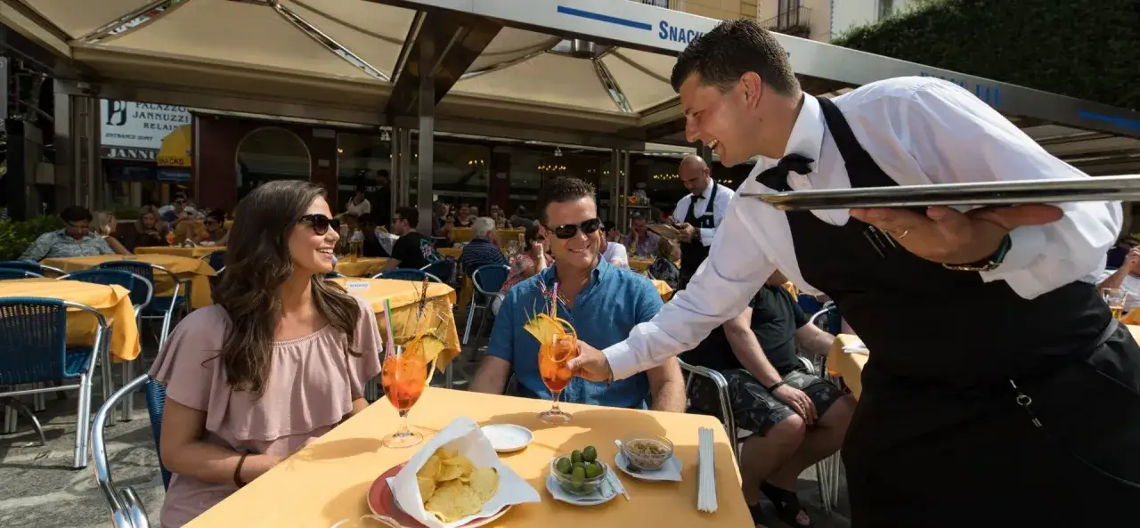 En servit&ouml;r serverar drinkar till ett leende par p&aring; en solig uteservering i Italien.