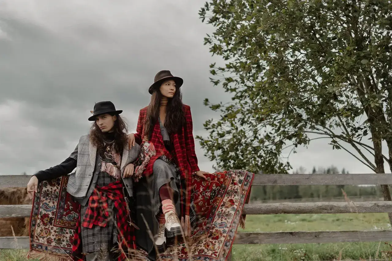Dos modelos con sombreros y abrigos a cuadros, sentados en una valla de madera, envueltos en alfombras con un marcado estilo bohemio.