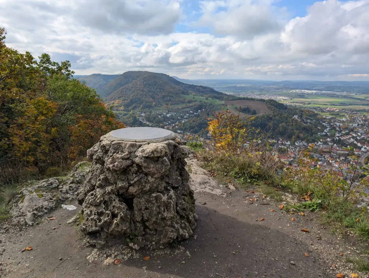 Blick vom Gipfelkreuz auf Heubach. Ein steinernes Monument mit einer Metallplatte steht im Vordergrund, während sich im Tal die Stadt und bewaldete Hügel erstrecken. Perfekt für alle, die gerne heubach wandern.