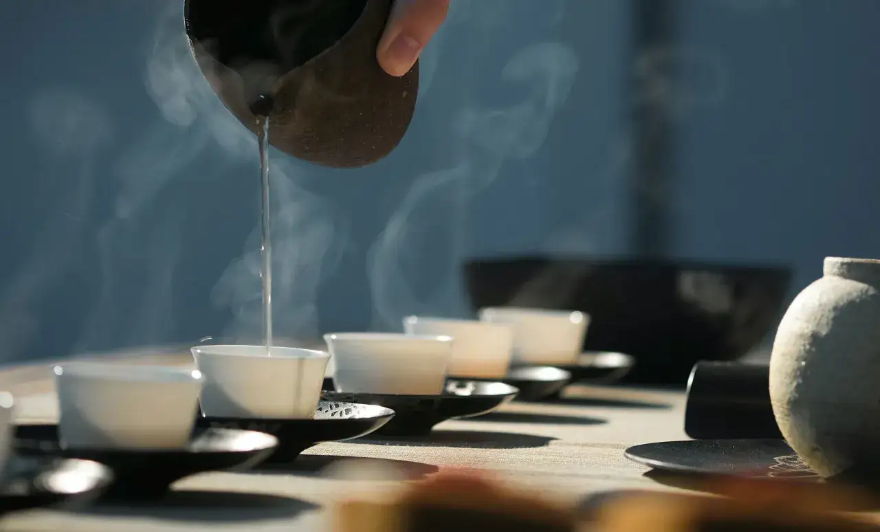 Service du thé : une main verse de l'eau chaude d'une tasse en terre cuite dans de petites tasses blanches alignées, la vapeur s'élevant dans l'air. Le noir des soucoupes contraste avec la blancheur des tasses.