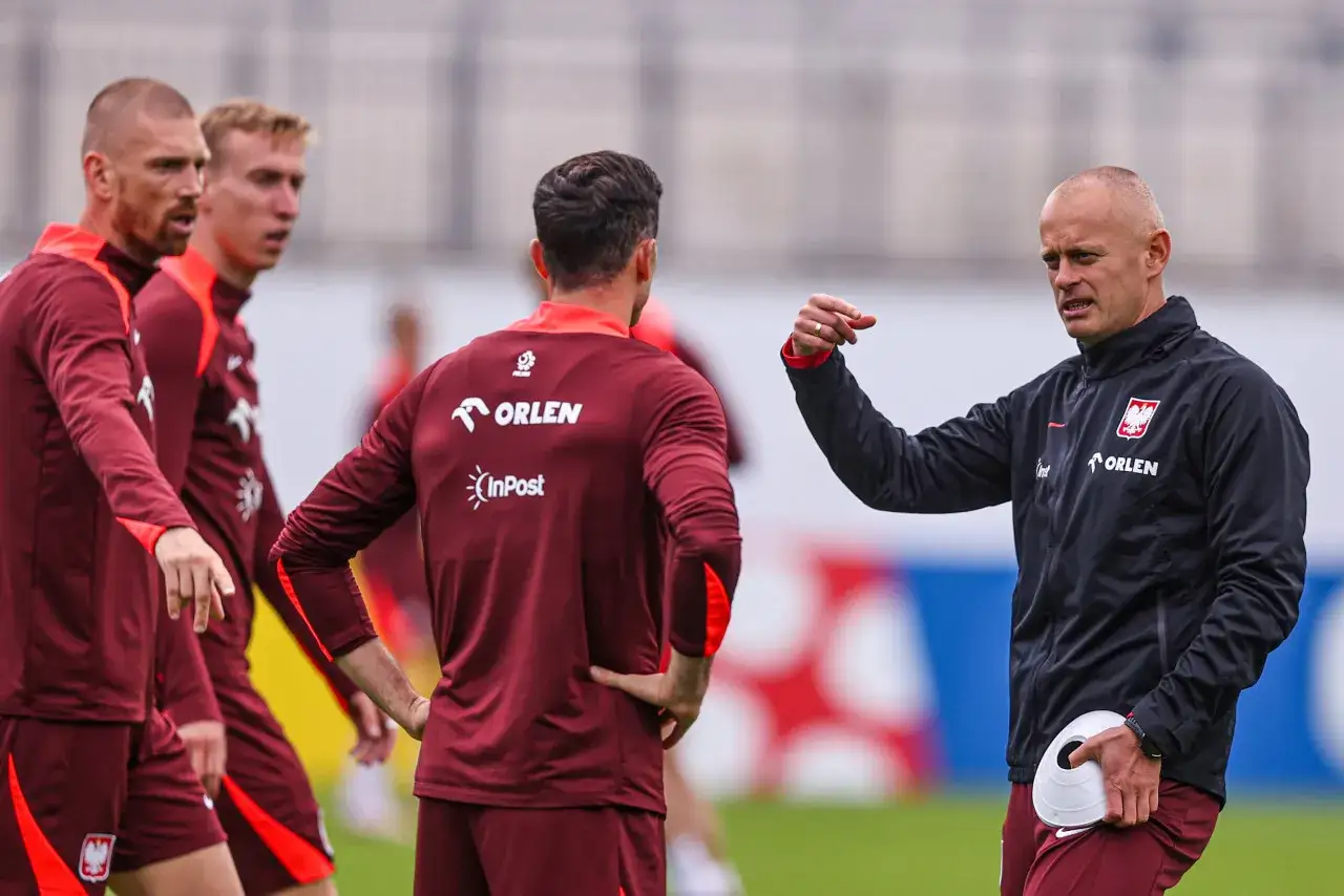 Marcin Włodarski trener, w czarnej kurtce z orłem, instruuje piłkarzy w bordowych strojach na boisku.