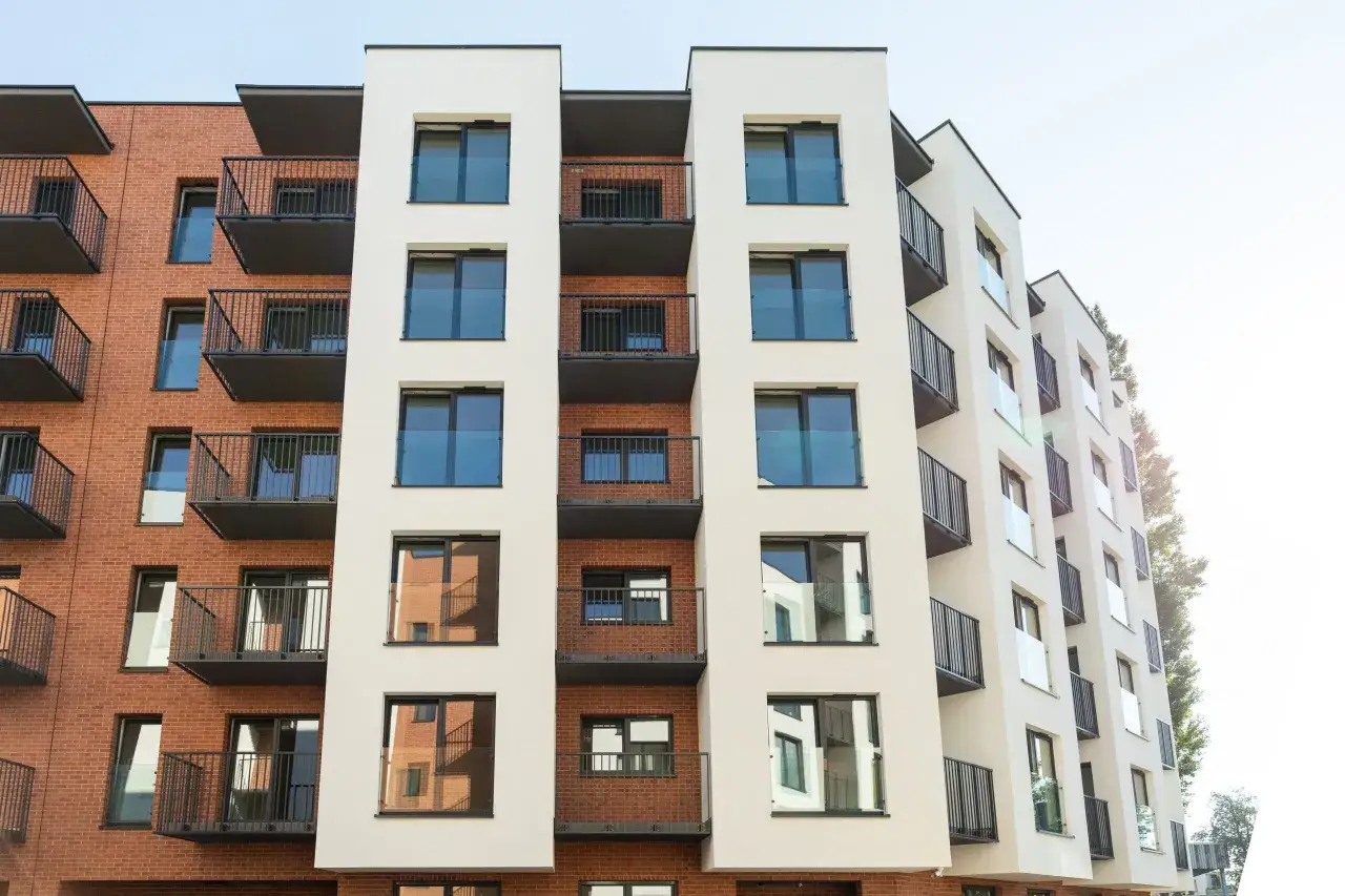 Nowoczesny budynek mieszkalny z ceglaną i jasną elewacją, liczne balkony z czarnymi balustradami. Idealne lokum salsa dla miłośnik&oacute;w miejskiego stylu.