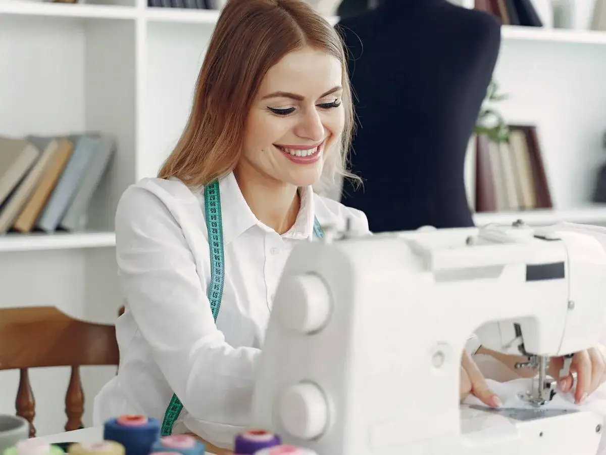 Mujer sonriente cosiendo ropa en m&aacute;quina de coser, con cinta m&eacute;trica al cuello.