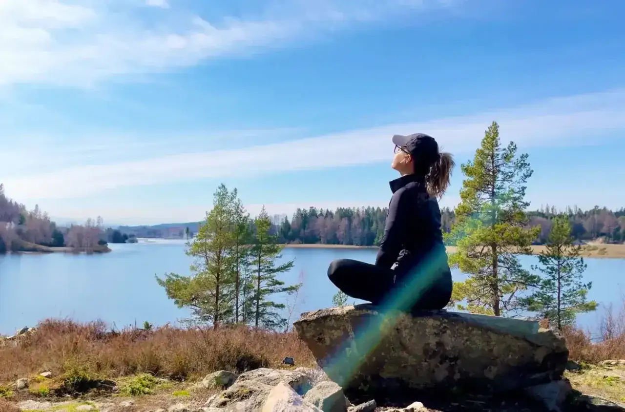 Kvinna sitter på en sten vid en sjö och samlar mental styrka. Solen lyser och himlen är blå.