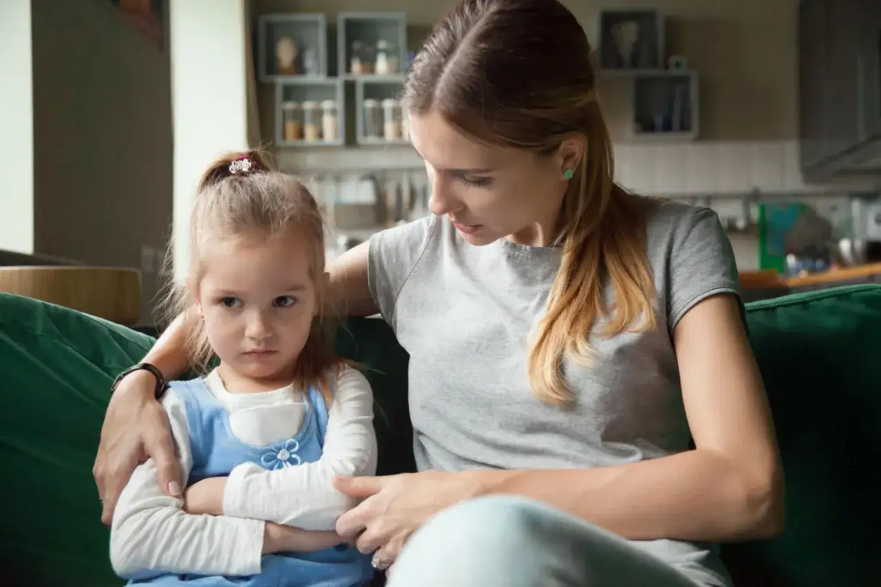 Mama próbuje uspokoić swoje 2-letnie dziecko, które przeżywa histerię. Dziewczynka ma skrzyżowane ręce i obrażoną minę.