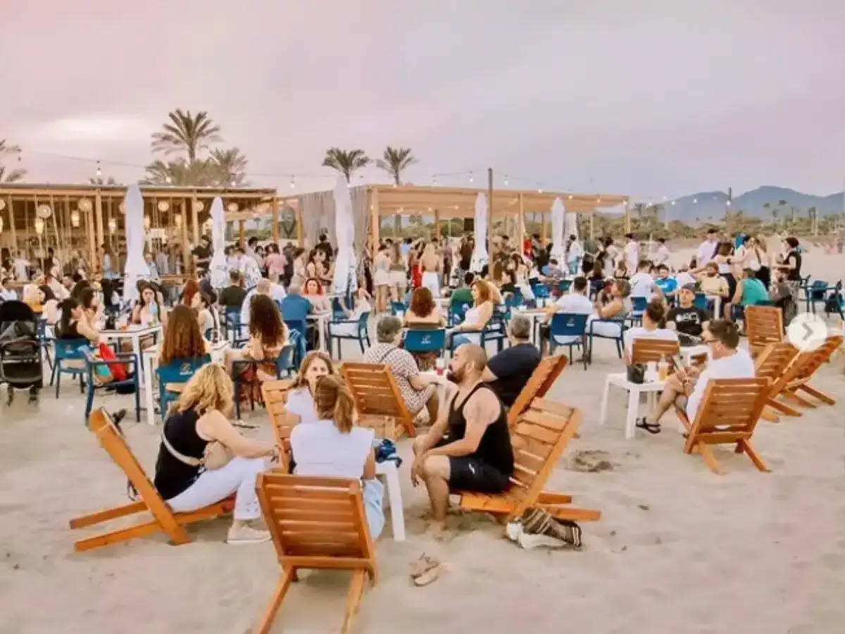 Gente disfrutando en un **som festival** playero, con mesas, sillas y palmeras bajo un cielo crepuscular.