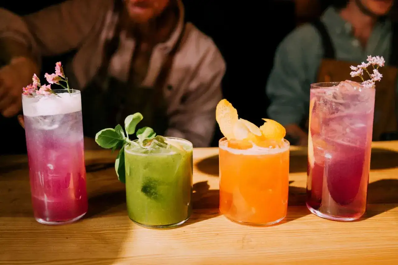 Quatre cocktails colorés, dont un cocktail 5 alcool, sont présentés sur un bar en bois. L'un est rose avec des fleurs, un autre vert avec des feuilles, un troisième orange avec des zestes, et le dernier violet.