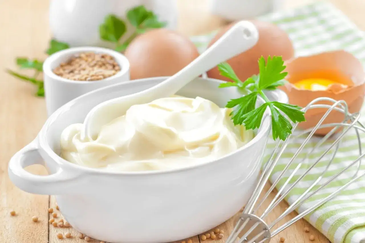 Un bol de mayonnaise maison sans œuf, garni de persil, avec des œufs, de la moutarde et un fouet à proximité.
