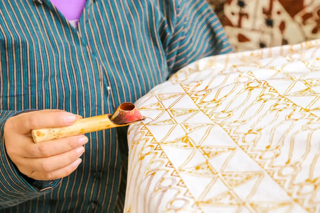 Eine Hand h&auml;lt ein Werkzeug, um mit Wachs Muster auf Stoff zu malen. Traditionelle Batik Techniken werden angewendet, um ein kunstvolles Design zu schaffen.