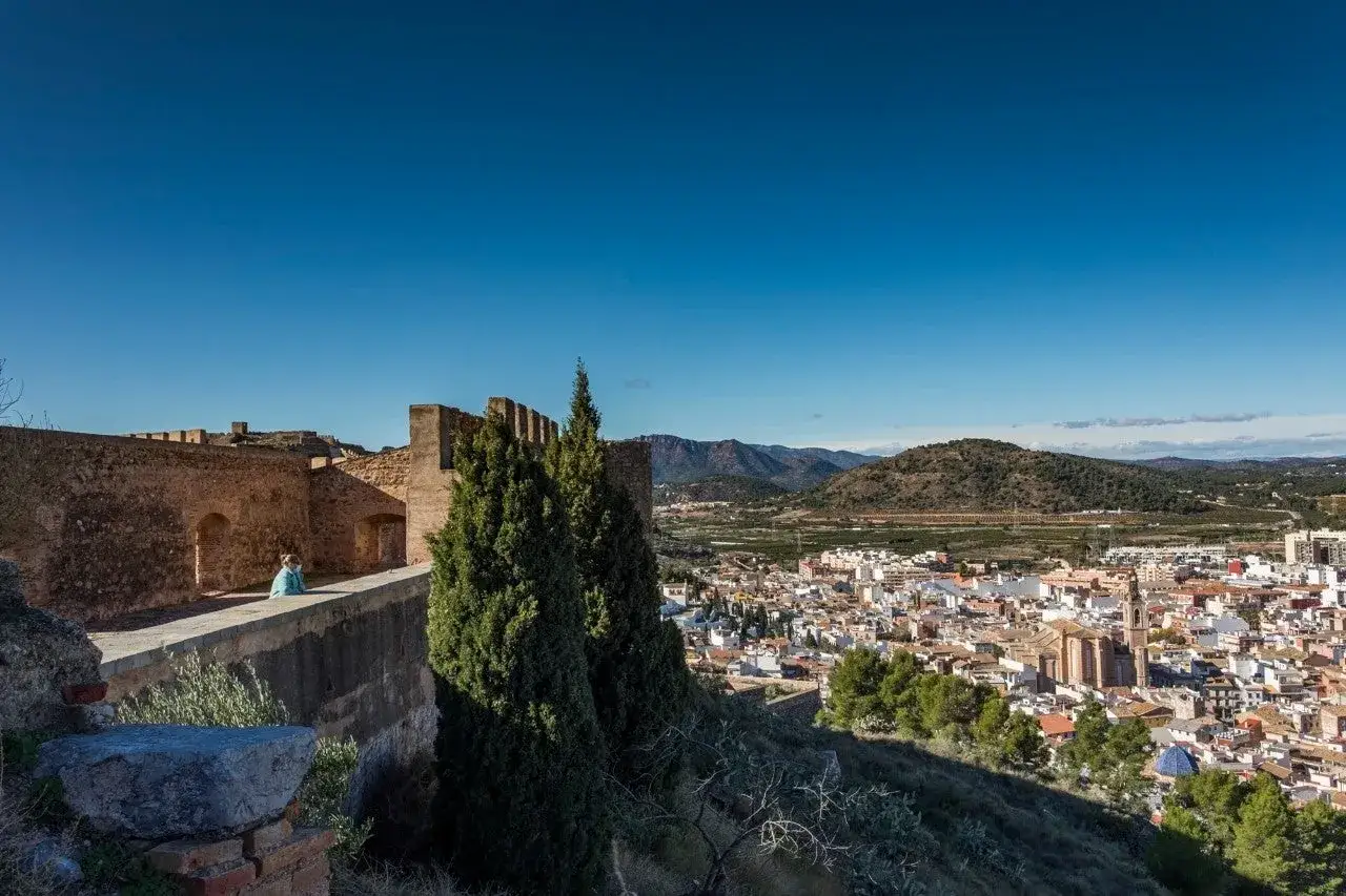 Vista panor&aacute;mica del **castillo de Sagunto** con una persona contemplando el paisaje urbano y las monta&ntilde;as al fondo.