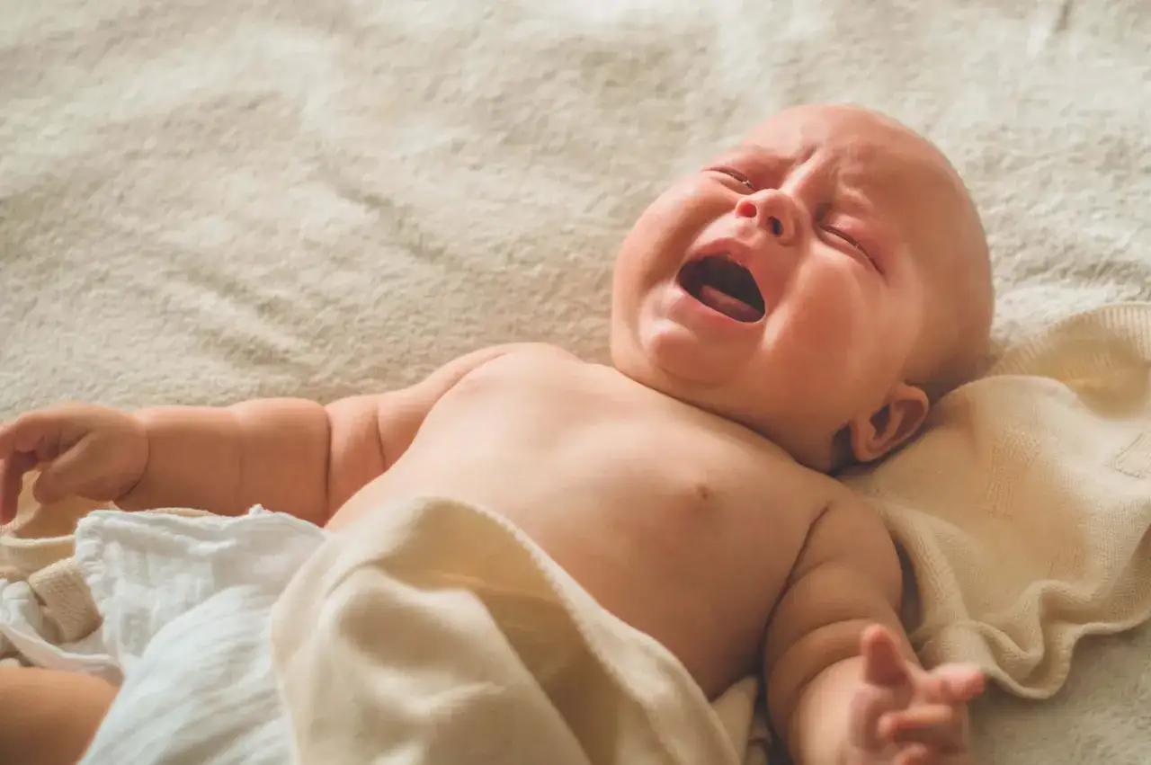 Un neonato piange disperatamente, con gli occhi chiusi e la bocca spalancata. Le sue manine sono tese, come a cercare conforto.