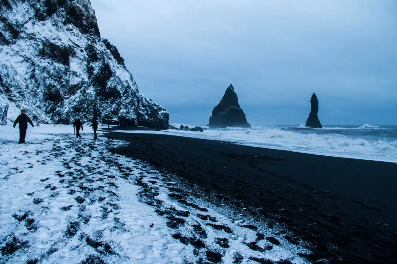 Islandia, listopad. Śnieg na czarnej plaży Reynisfjara, ludzie spacerują przy skalnych formacjach w morzu.