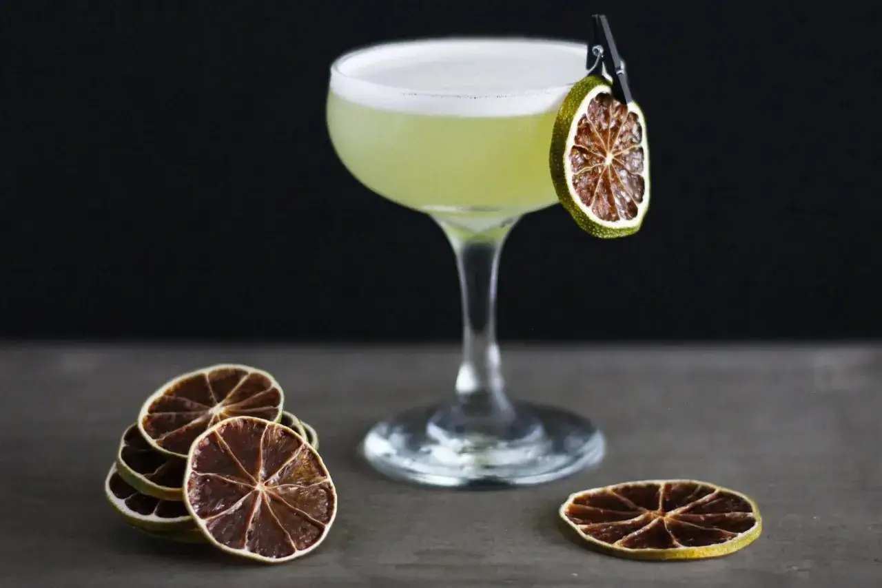 Zielony, orzeźwiający drink z pianką, ozdobiony suszoną limonką na klipsie. Obok stosik plasterk&oacute;w cytrus&oacute;w. Idealny na letni wiecz&oacute;r, ostatnie słowo cocktail.