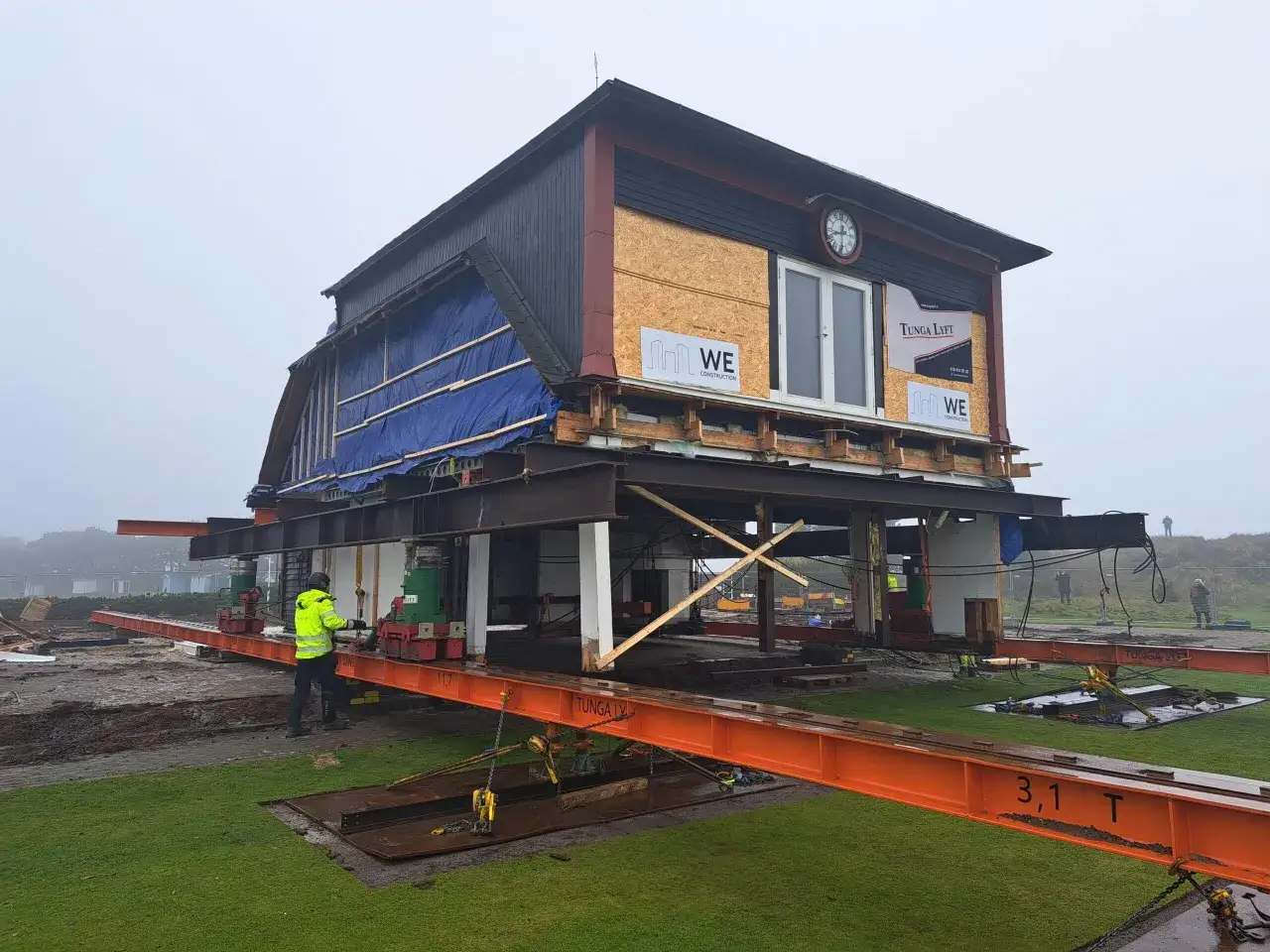 Ett hus lyfts på orange balkar för att flytta friggebod. En person i gul jacka övervakar arbetet.