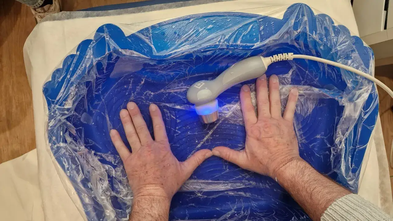 Mani immerse in un bagno blu per ultrasuoni in acqua per piedi, con un manipolo che emette luce blu.