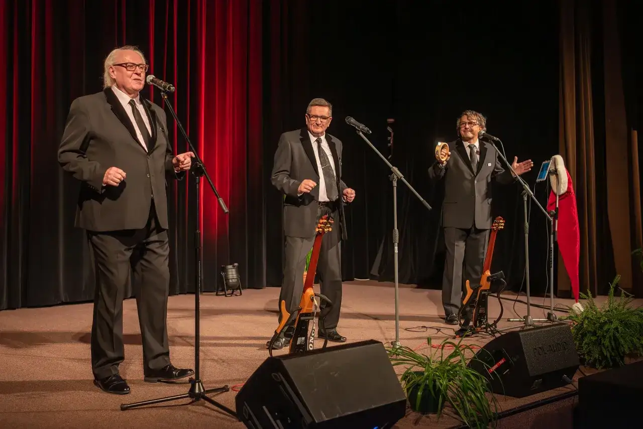 Trzech panów w garniturach, jak kabaret Otto, na scenie. Jeden gra na tamburynie, drugi trzyma gitarę, trzeci śpiewa.