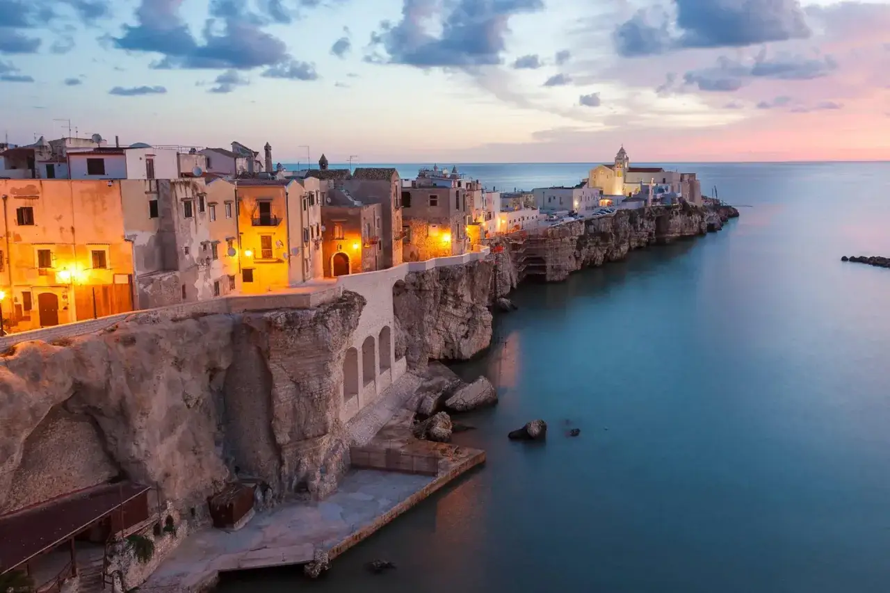 Piazzetta Petrone a Vieste, con case illuminate su scogliere a picco sul mare al tramonto.