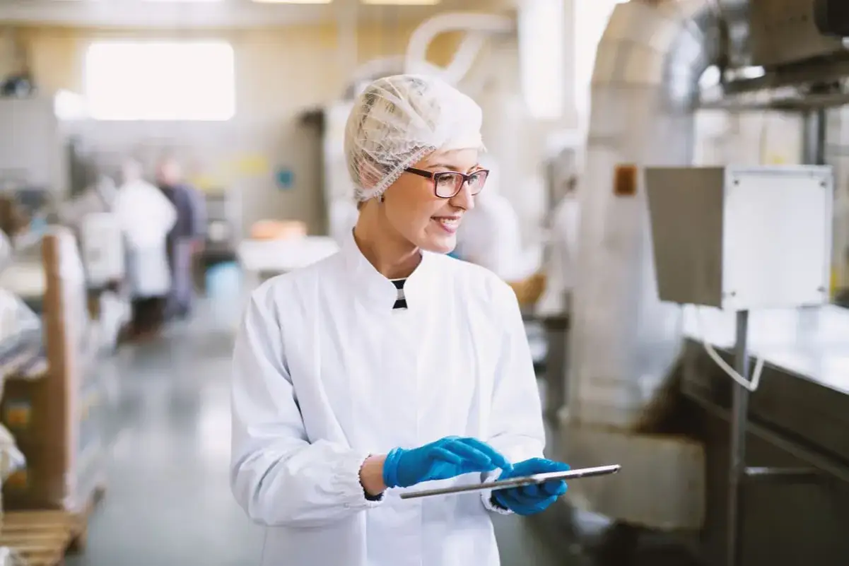Employée en blouse blanche et charlotte, effectuant un contrôle d'hygiène sur tablette dans une usine.