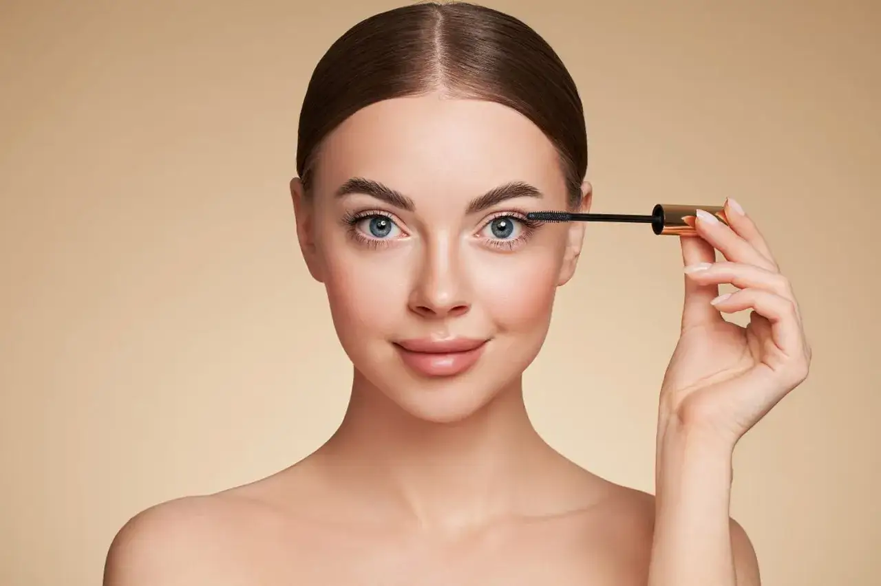 Une femme applique du mascara pour un maquillage des yeux ronds et globuleux, accentuant son regard bleu intense.