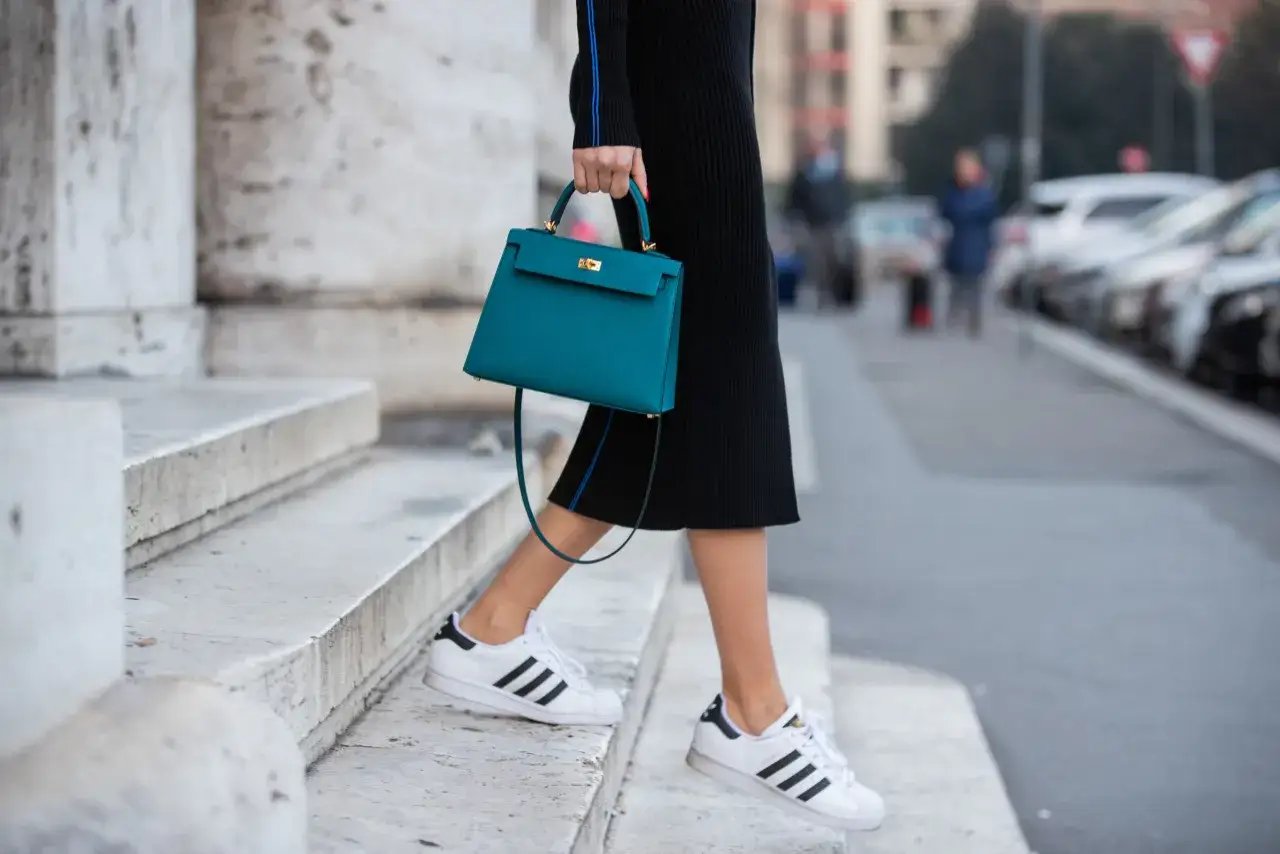 Donna con tubino nero, borsa turchese e scarpe basse Adidas bianche con strisce nere, scende le scale.