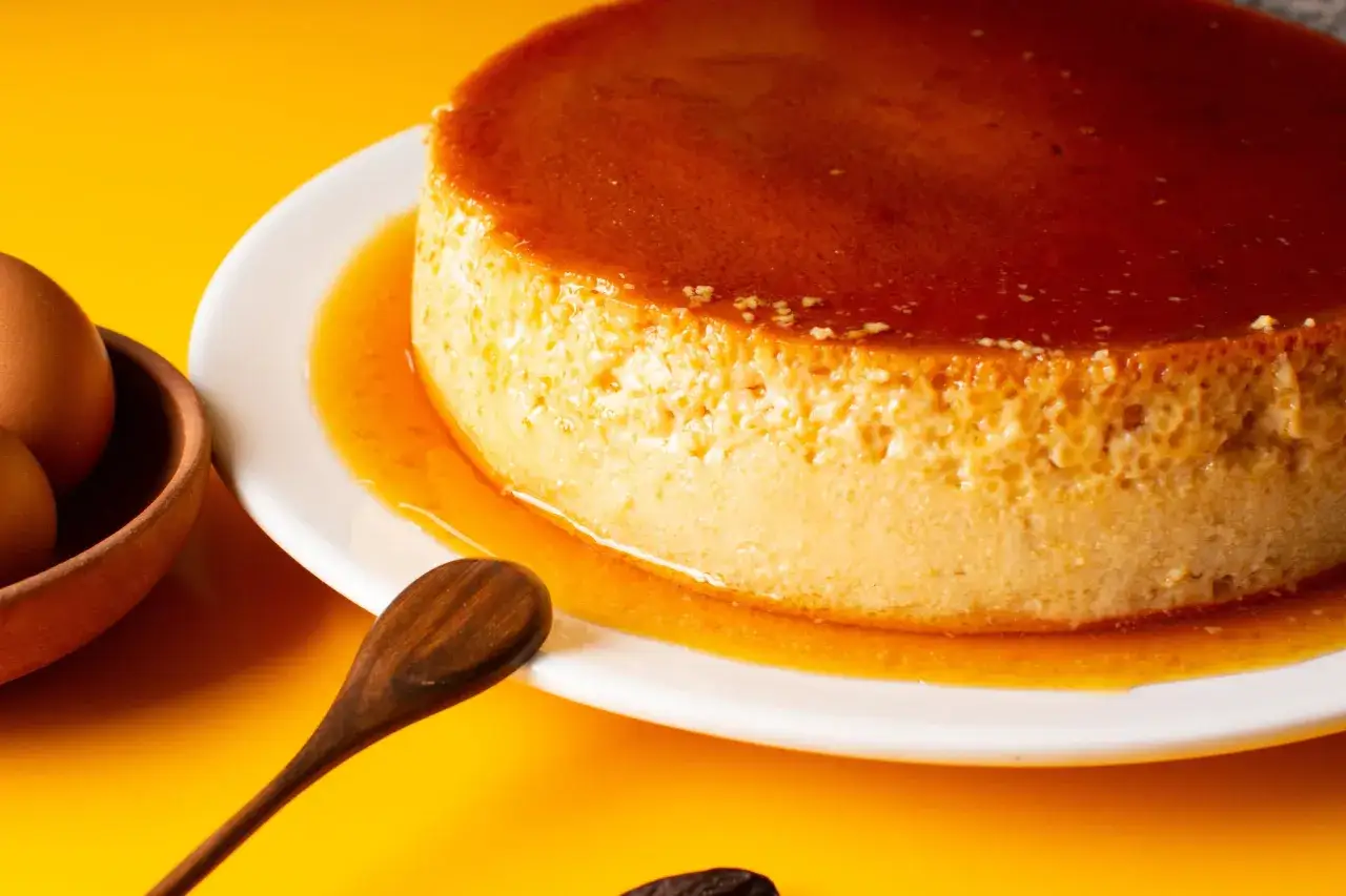 Un délicieux flan aux œufs, comme la recette flan aux œufs de nos grand-mères, baignant dans son caramel doré. Des œufs bruns dans un bol en terre cuite et une cuillère en bois complètent la scène.