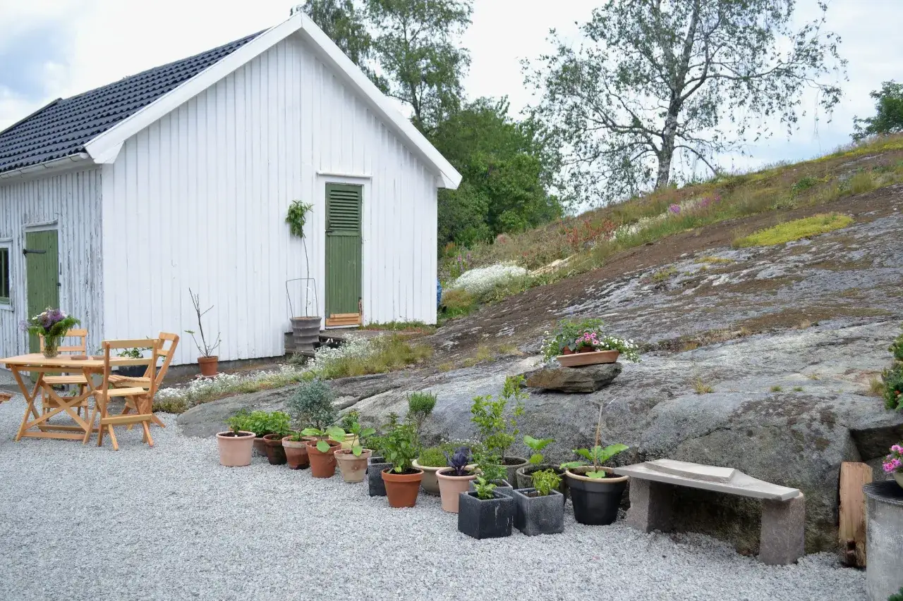 Vitt hus med gr&ouml;na detaljer och en uteplats med grus till uppfart. Tr&auml;dg&aring;rdsm&ouml;bler och krukor med v&auml;xter.