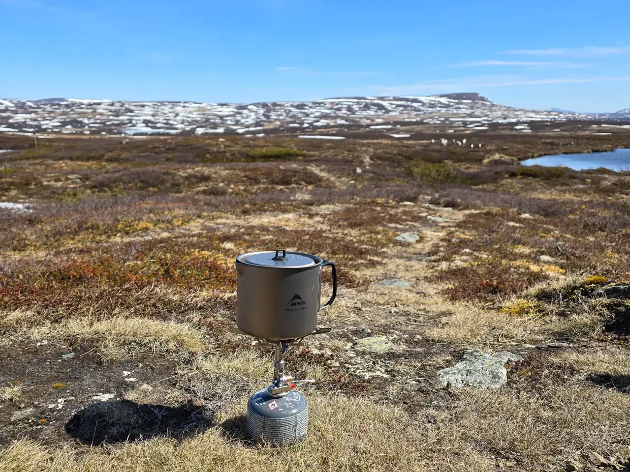 En kastrull på en campingkök i fjällandskap, redo för matlagning under en vandring. Snö och en sjö syns i bakgrunden.