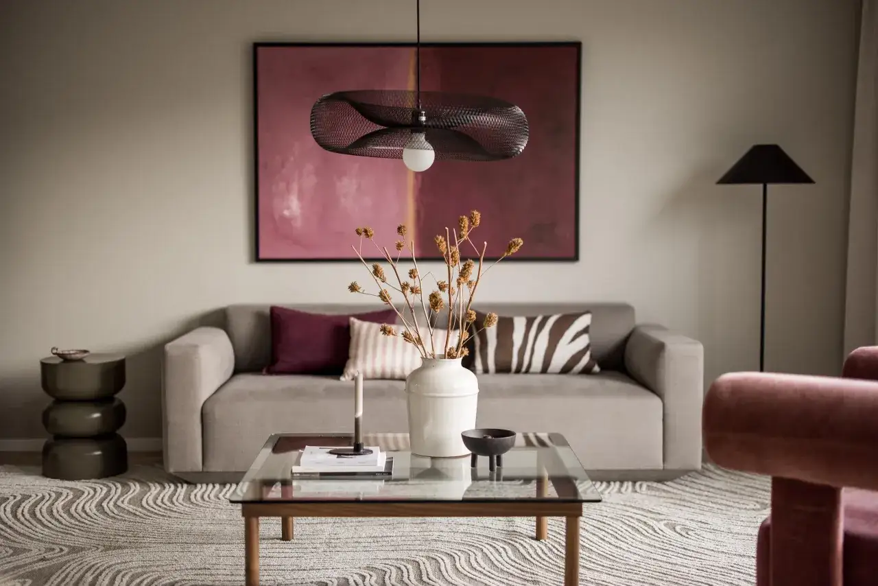 Ett mysigt tv-rum med en gr&aring; soffa, en stor tavla med rosa toner och en modern taklampa. Ett soffbord med en vas och torkade blommor skapar en inbjudande atmosf&auml;r.