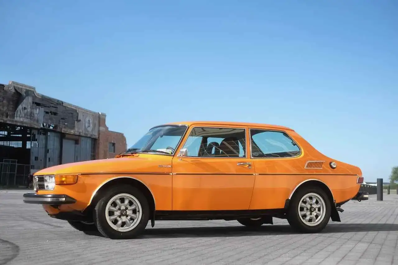 En orange Saab 99 från 70-talet står parkerad framför en gammal industribyggnad.