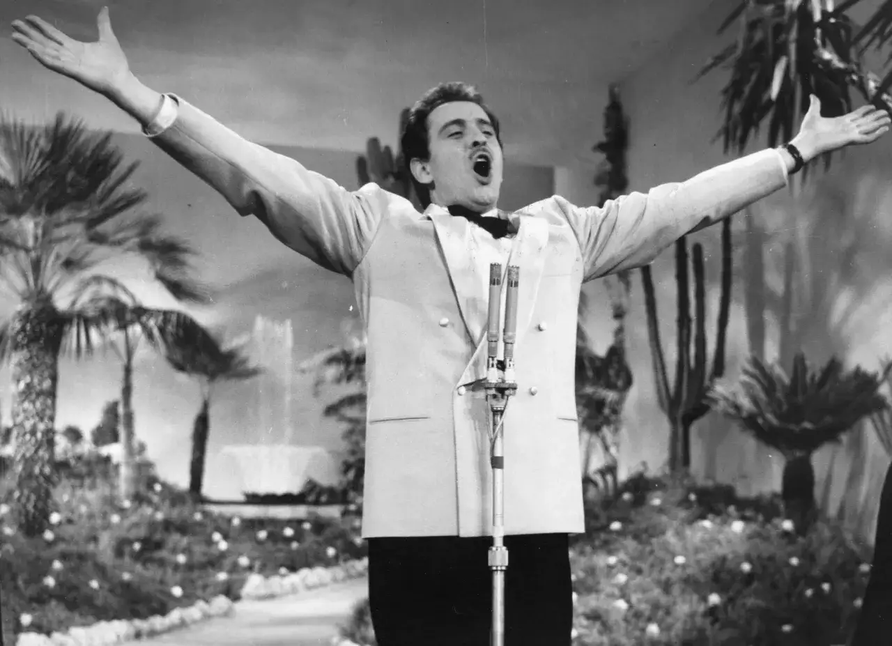 Domenico Modugno, con le braccia aperte, canta con passione su un palco decorato con piante esotiche.