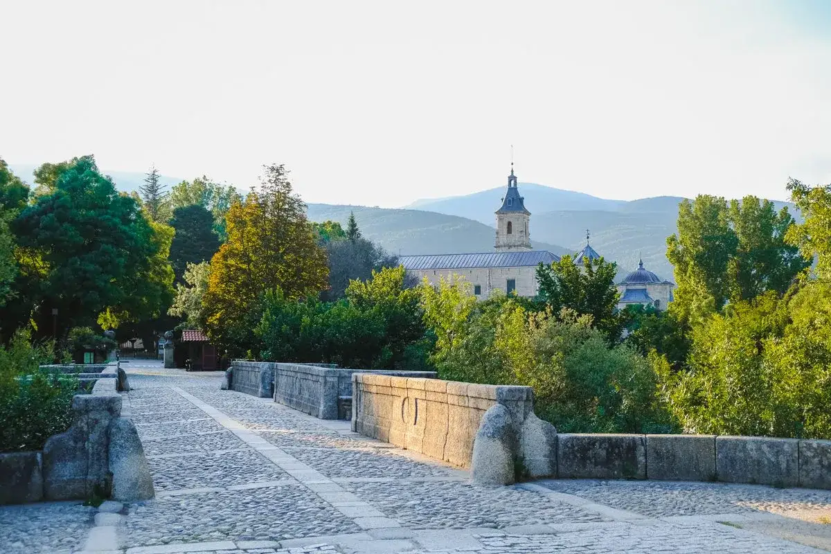Puente de piedra que conduce al monasterio del Paular, rodeado de &aacute;rboles frondosos y monta&ntilde;as al fondo.
