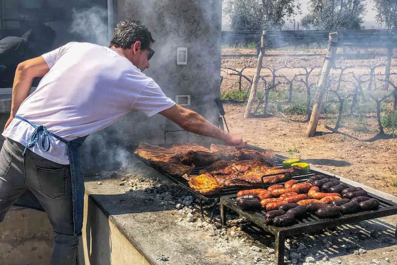 Un homme prépare un barbecue bois ou charbon, avec des côtes et des saucisses qui fument.