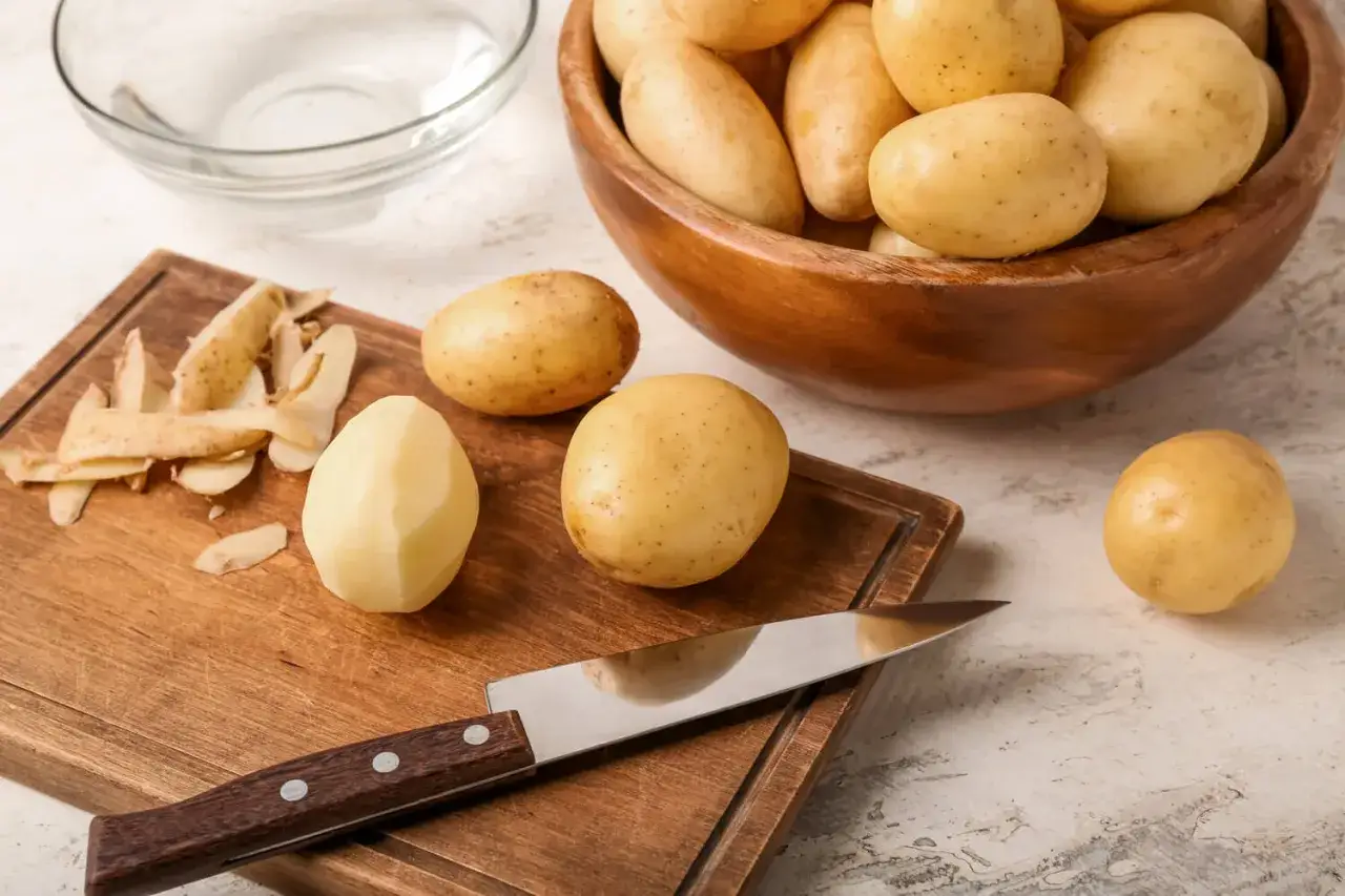 Einige Kartoffeln werden gesch&auml;lt, eine liegt bereits gesch&auml;lt auf einem Holzbrett neben einem Messer. Eine Sch&uuml;ssel mit Kartoffeln steht im Hintergrund. Die Frage 