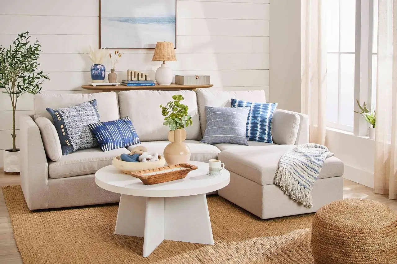 Gem&uuml;tliches Wohnzimmer im New England Style mit hellem Sofa, Kissen in Blau-Wei&szlig;-T&ouml;nen, einem runden Couchtisch und Jute-Teppich.