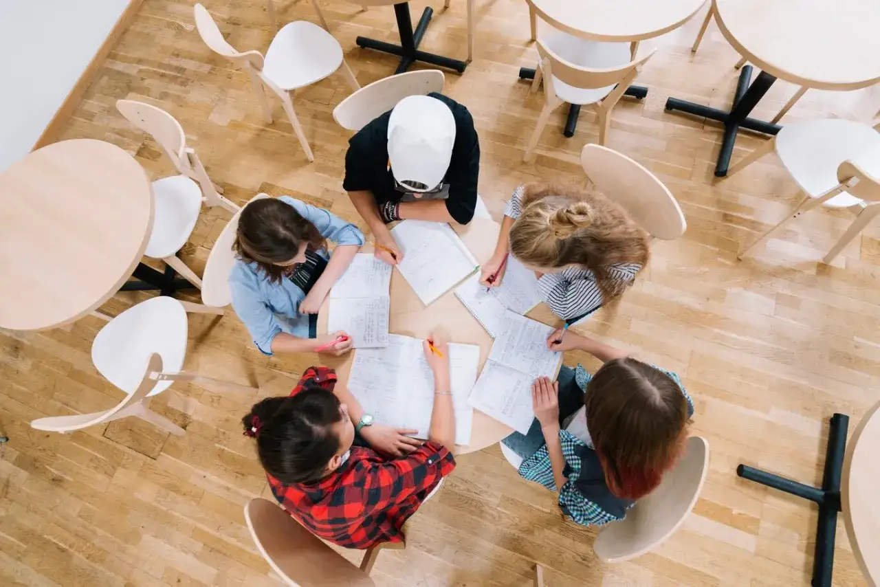 Cinco j&oacute;venes participan en din&aacute;micas de grupo, sentados alrededor de una mesa redonda, escribiendo y colaborando en sus estudios.