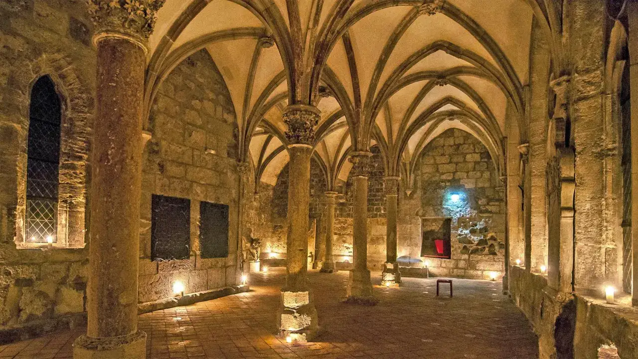 Gewölbter Raum mit Säulen, der an ein mittelalterliches Kloster erinnert. Kerzenlicht beleuchtet die steinernen Wände und den Boden.