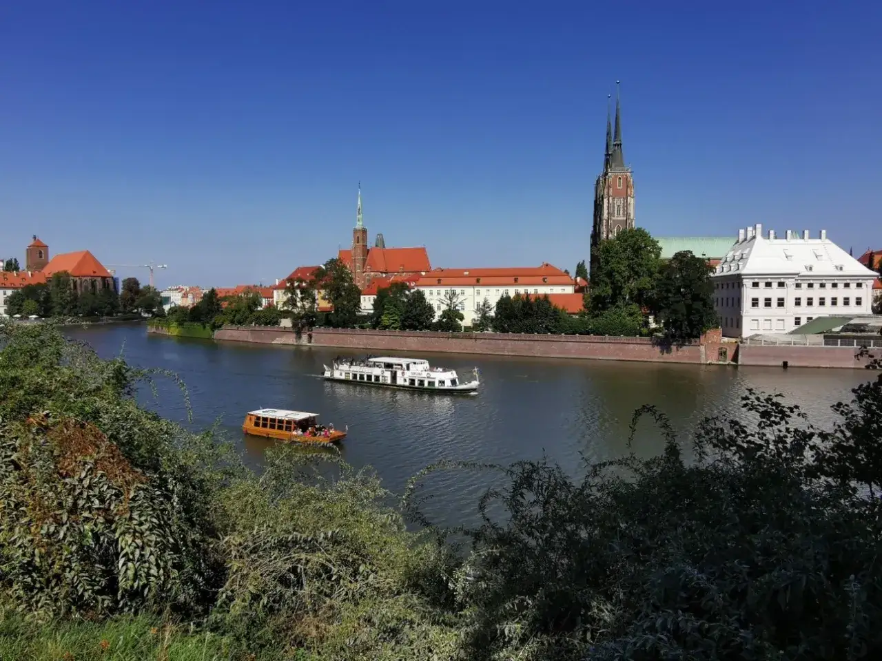 Wrocławskie bulwary nad Odrą, idealne miejsce, gdzie na spacer we Wrocławiu. Dwa statki płyną po rzece, a w tle widać zabytkowe budynki.