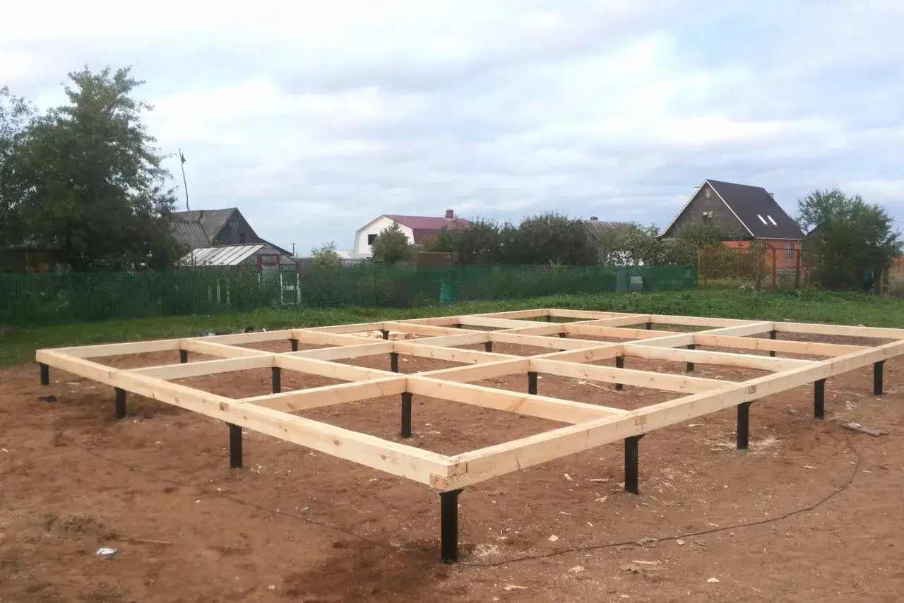Drewniana konstrukcja na słupach, przygotowywana jako fundament słupowy pod przyszły dom. W tle widać zabudowania i drzewa.