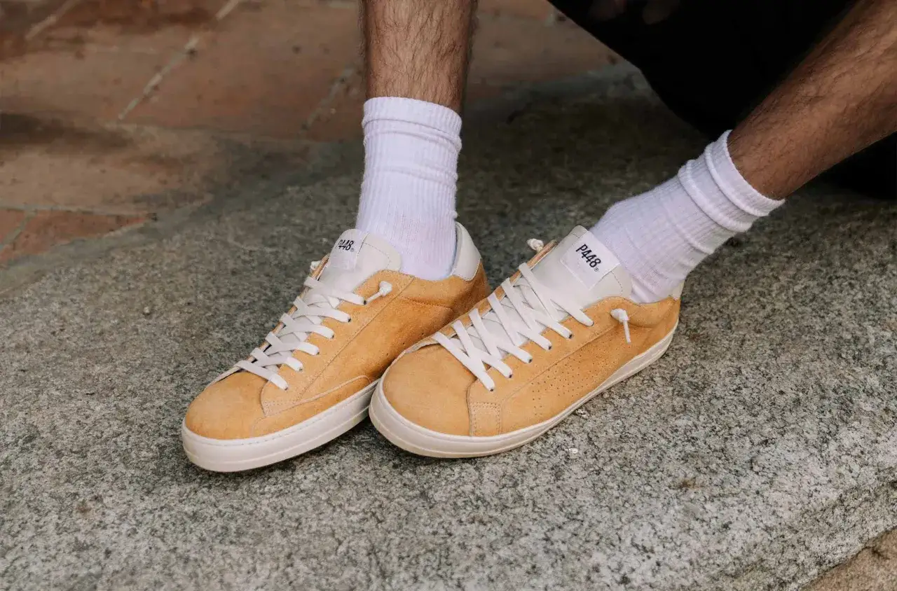 Scarpe P448 uomo scamosciate color ocra con lacci bianchi e calzini bianchi, appoggiate su un fondo di pietra grezza.