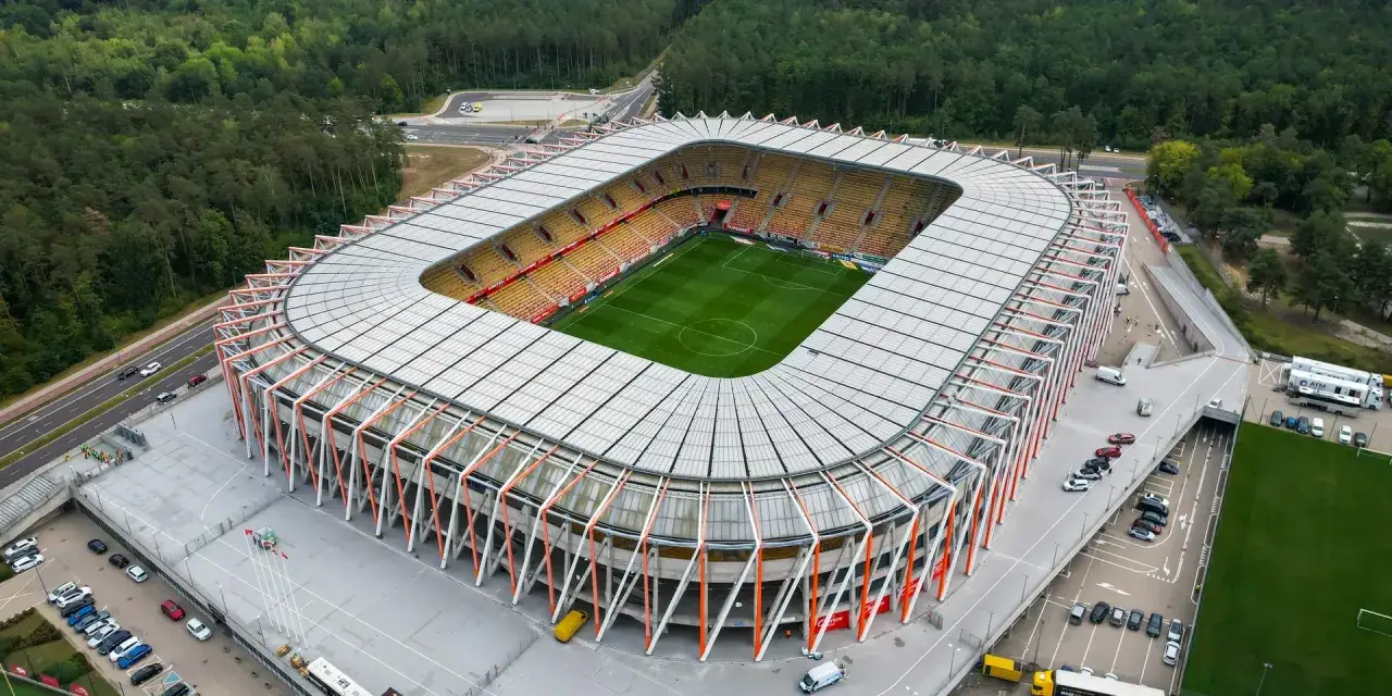 Nowoczesny stadion piłkarski, gotowy na emocjonujące rozgrywki ekstraklasy. Otoczony lasem, z zieloną murawą i ż&oacute;łtymi trybunami.