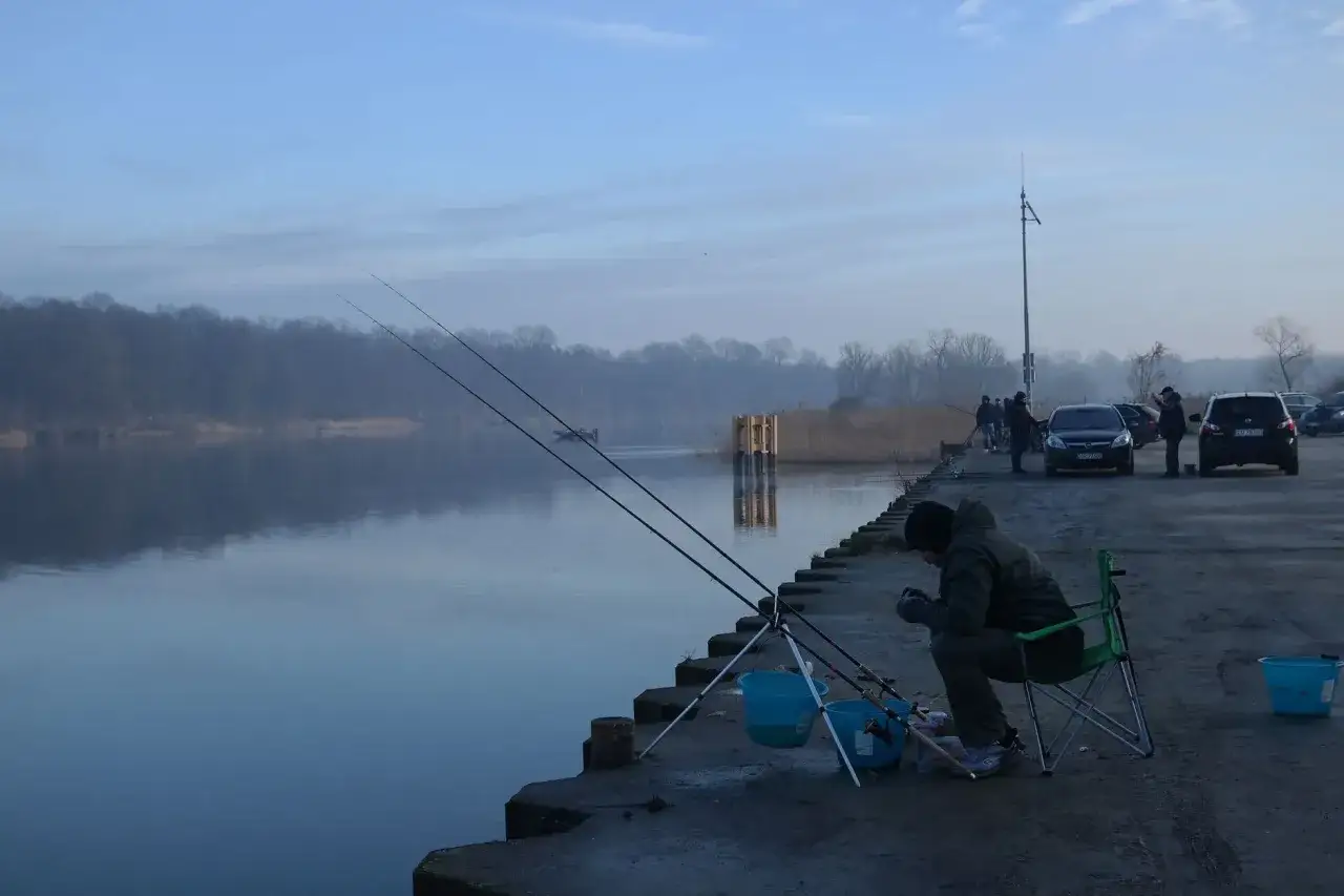 Wczesne godziny brania ryb. Wędkarz siedzi na krześle nad wodą, czekając na branie. W tle widać inne osoby łowiące ryby i zaparkowane samochody.