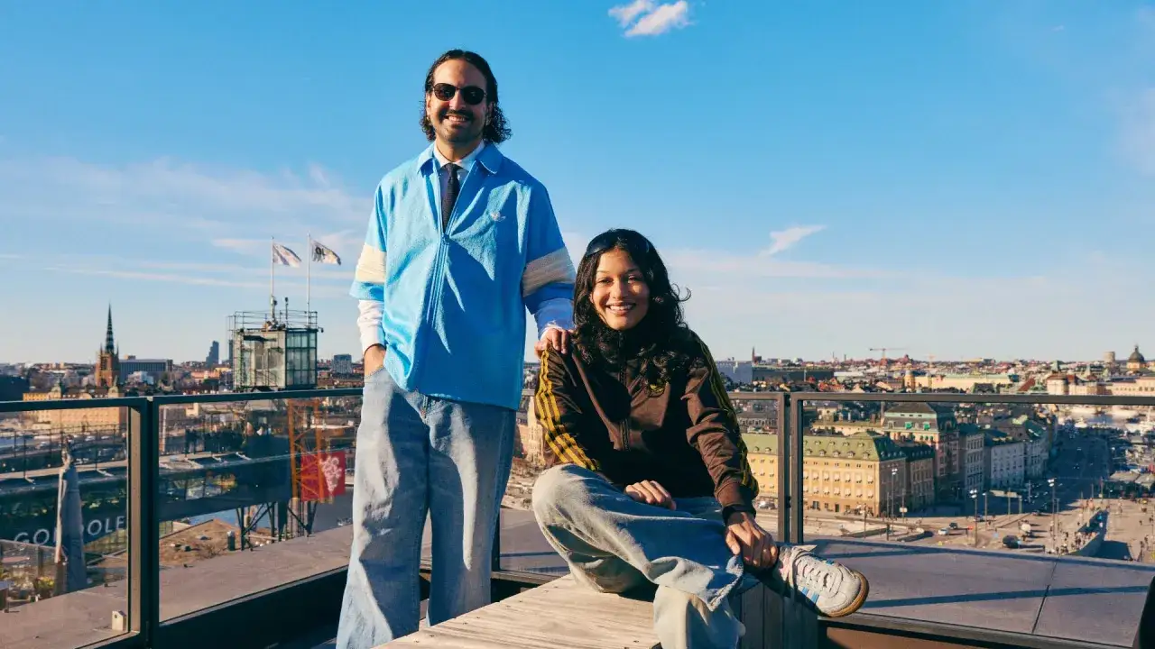 Två glada personer på en takterrass med utsikt över Stockholm, en solig dag. I bakgrunden syns stadens silhuett och en glimt av pelago slussen.